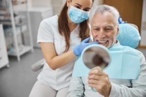 an older man smiling in the mirror at his new dental implants with his dentist